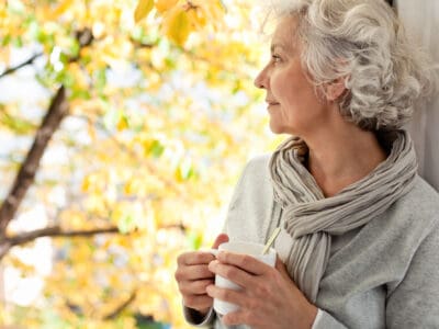 Woman with hearing loss looks out of window, holding a cup of coffee and thinking about taking a free online hearing test on the Lexie Hearing website.