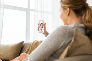 Woman sitting on couch, wearing an invisible hearing aid, consults with hearing health practitioner and hearing aid expert on her smartphone, explaining what is teleaudiology through a smartphone.