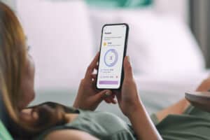 Woman lies on her couch looking at her mobile phone. On the screen is the Lexie Rewards program she has access to throuh her Lexie Lumen best value hearing aid