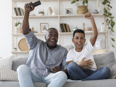Grandfather and grandson sitting on the couch cheering after something happened on the television. The grandfather with hearing loss is using assistive devices such as hearing aids to better hear the TV.