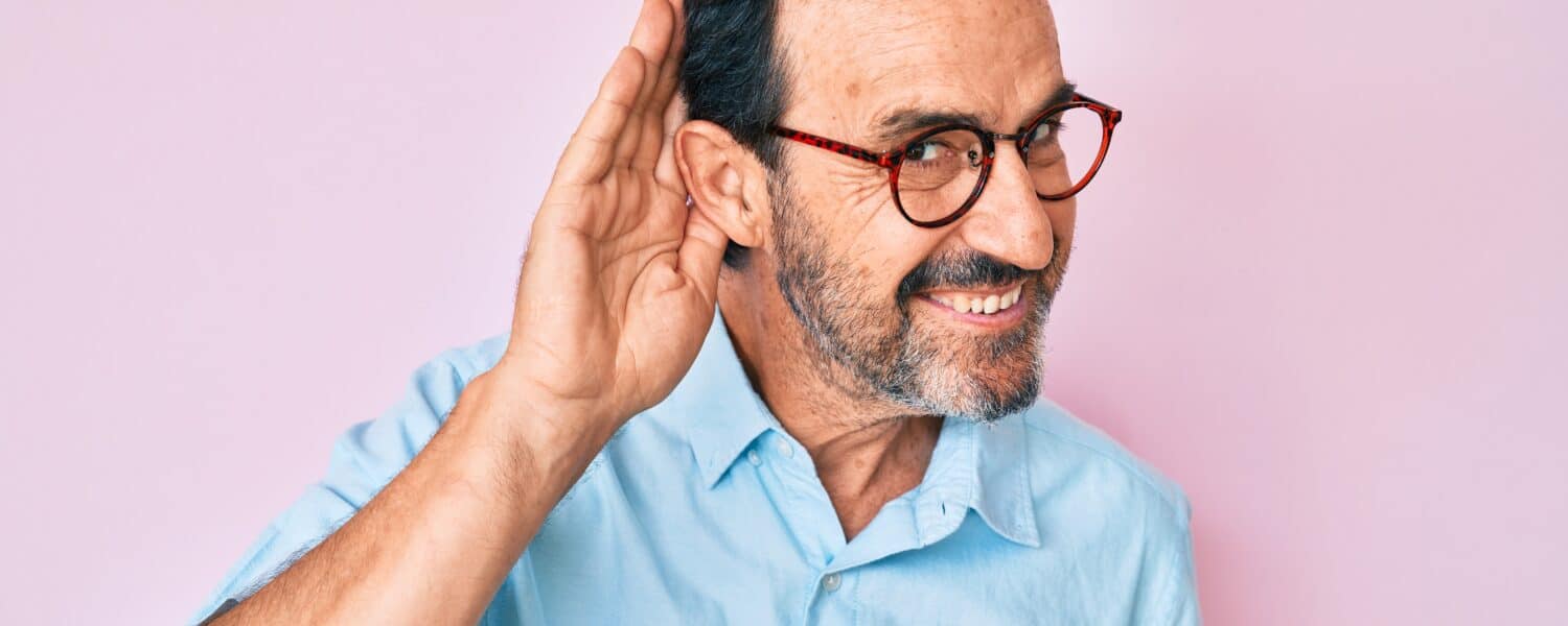 Man smiling and holding is ear asif he has one-sided hearing loss. He is considering getting invisible hearing aids.