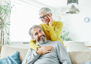 elderly couple, woman embracing the man from the back while he sits on the couch. The woman holds her ear asif she has a hearing loss in it.