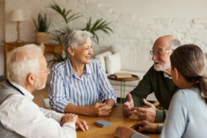 Group of elderly friends sitting around a table playing cards and talking about Lexie Lumen hearing aids. The man asks 